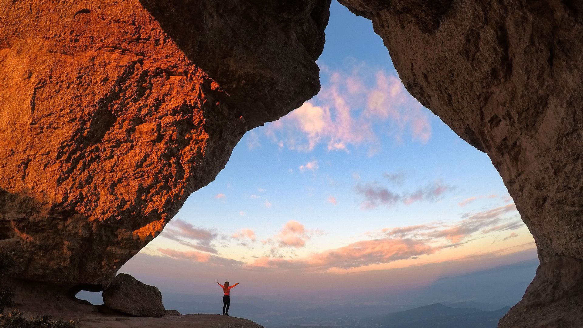 Imagem de uma pessoa de braços abertos olhando por uma grande abertura entre pedras para o por do sol